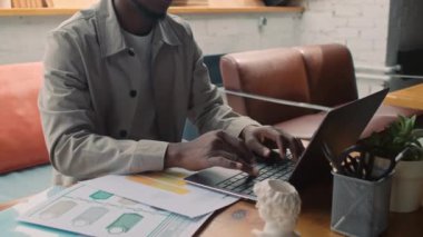 Medium close-up shot of hands and midsection of anonymous African American male freelancer in casual clothes sitting at table on couch in coworking office and typing on laptop