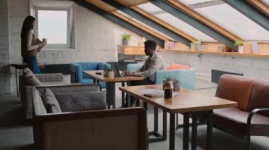 Full zoom shot of African American man sitting at table in loft coworking office, typing on laptop, and female Asian colleague or manager with documents walking in, sitting down for business meeting