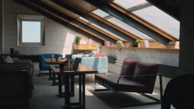 Full no people shot of interior of coworking facility with white brick walls, windows on sloped roof, laptop and stationery on tables, couches, and books on windowsill, in daytime