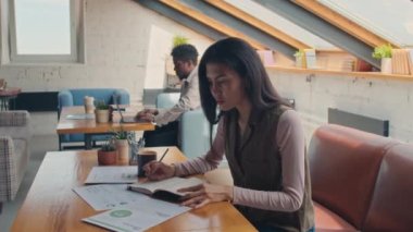 Medium shot of multiracial customers sitting at tables in coworking facility - African American man typing on laptop, and busy Asian female student reading presentation and making notes in workbook