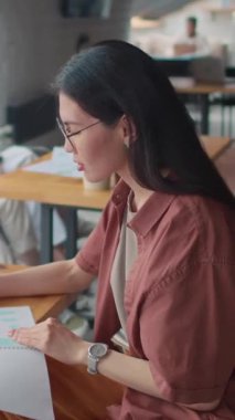 Genç Asyalı gözlüklü kız öğrencinin orta boy dikey portresi. Çalışma masasında ya da kantinde oturuyor, sunum ya da ders notları okuyor, sonra kameraya bakıyor ve gülümsüyor.