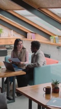 Genç Asyalı kadın ve Afrikalı Amerikalı erkek partnerin tam dikey çekimleri tavan arası çalışma ofisinde koltukta otururken, dizüstü bilgisayarı masada tutarken ve iş teklifini tartışırken.