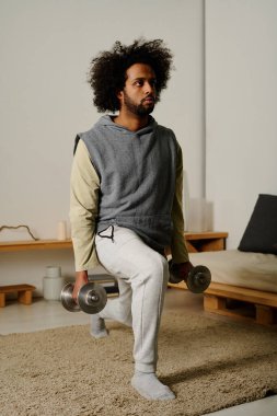 Elinde ağır metal dambıllar olan aktif bir adam yatak odasının zemininde duruyor ve egzersiz sırasında bacak esneme egzersizi yapıyor.