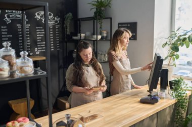 Zihinsel engelli genç bir kadın bilgisayar ekranını işaret eden meslektaşının yanında durup not defterindeki notlara bakıyor.