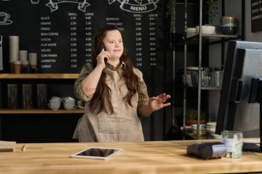 Zihinsel engelli genç bir kadın bilgisayarın karşısında duruyor ve kahve müşterileriyle cep telefonuyla iletişim kuruyor.
