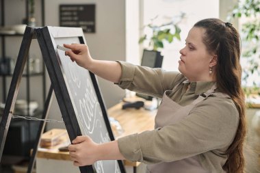 Kafe 'nin zihinsel engelli genç bayan çalışanı kameranın önünde dururken menüyü tahtaya beyaz tebeşirle yazıyor.