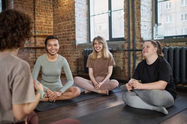 Kültürler arası etkin üç genç kadın sohbet sırasında spor hocalarına bakıyor ve paspaslarda oturuyor ve tatilin tadını çıkarıyor.
