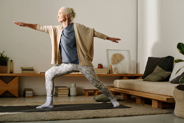 Elderly active woman in casualwear keeping arms and legs outstretched while standing on mat in bedroom and doing yoga exercise