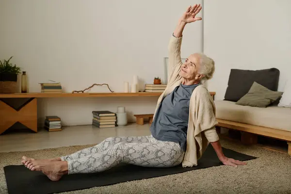 Retired woman in activewear sitting on mat on the floor of bedroom and doing side bends with raised arm while working out after sleep