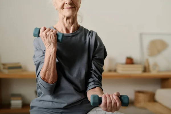 Active retired woman with dumbbells in hands pumping arm muscles during morning workout while sitting in front of camera