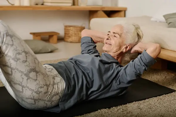 Senior active woman in leggins and grey cotton pullover doing sit ups on black mat during morning workout on the floor of bedroom