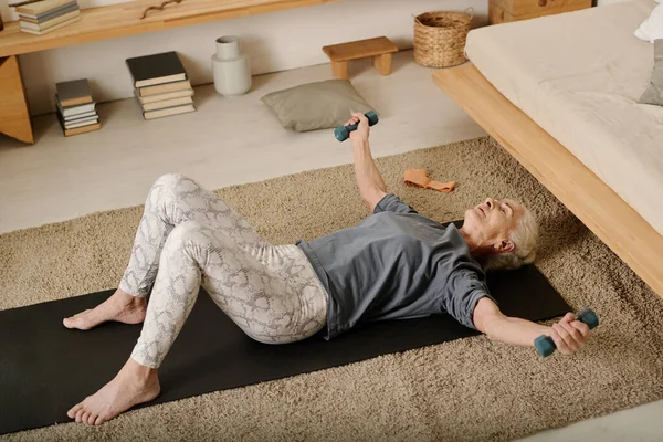 stock image High angle of aged woman in activewear lying on mat in bedroom and doing exercise for arm and chest muscles during morning workout