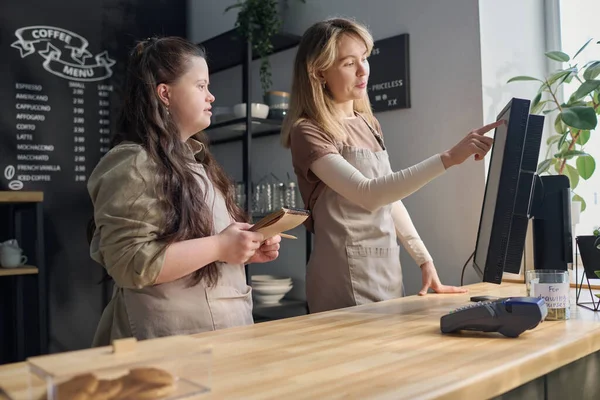 Young woman pointing at computer screen while explaining female colleague with disability her how to count cost of orders