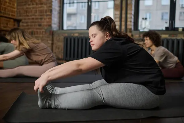 Side view of young active woman with Down syndrome sitting on mat in gym and stretching arms towards toes during physical exercise