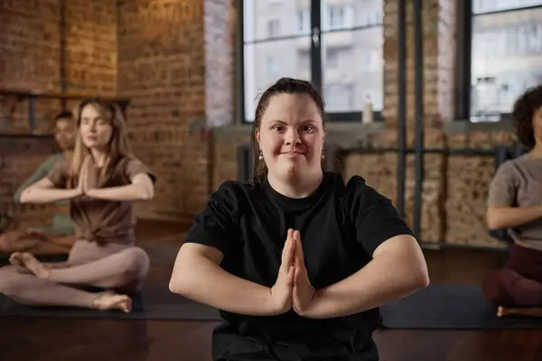 Active girl with disability keeping her hands put together against chest and looking at camera during yoga practice with other women
