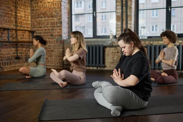 Female fitness trainer with disability keeping hands put together against chest while sitting on the floor and practicing yoga in gym