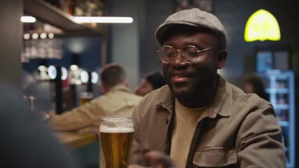 グラスとキャップで黒人の笑顔と話し クラブで夕方に一緒にビールをクランジングするという肩の映像 — ストック動画