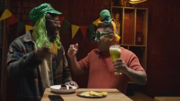 緑の帽子でアフリカ系アメリカ人と白人の男性のメディア映像は バーでSt Patricks Dayを祝っている間 緑のビールのパイントでテーブルの近くで踊るウィッグと — ストック動画