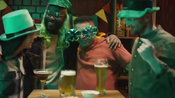 居酒屋のテーブルの近くで踊る緑の帽子をかぶる陽気な多民族の友人のメディア映像と 聖パトリックの日を祝いながらエメラルドラガーのメガネを凝視 — ストック動画