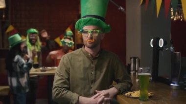 Beyaz adamın orta boy portresi. Büyük yeşil cüce cüce şapkası ve sahte bıyıklı komik gözlükler takıyor. Kutlama gününde İrlanda barında kameraya poz veriyor.