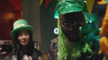 17 Mart 'ta barda Aziz Patrick Günü' nü kutlarken yeşil şapkalı ve cızırdayan bira bardakları takan çok ırklı yetişkin arkadaşların göğüs röntgeni.