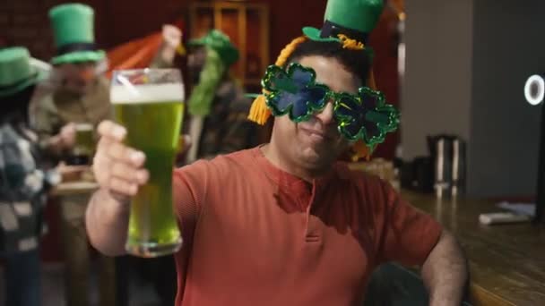 シャムロックグラスと小さな帽子で陽気な男の肖像画をチェストアップ セントパトリックスデーのバーでパーティー中にカメラを見ながら緑のビールと笑顔を提供 — ストック動画