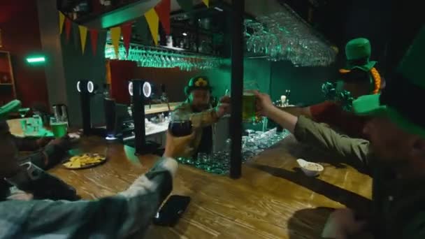 セントパトリックデーのバーカウンターで緑の帽子で友人と座ってバーテンダーと祝う緑とクラフトビールのクランキンググラス — ストック動画