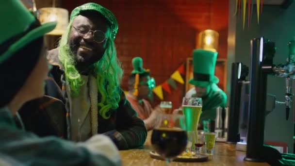緑の帽子でアフリカ系アメリカ人の男性のサイド映像と バーでセントパトリックの日のパーティーでビールと一緒にバーカウンターに座っている認識できない女性と話すウィッグ — ストック動画
