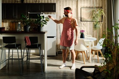 Önlüklü ve günlük giysili bir adam mutfak tezgahının yanında durup saksılarda yetişen ev bitkilerini sularken onlara bakıyor.