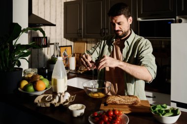 Tişörtlü genç adam mutfak tezgahında durup kahvaltıya omlet hazırlarken cam kaseye taze çiğ yumurta kırıyor.