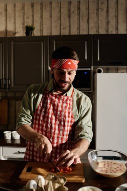 Saç bandı ve önlüklü adam mutfak masasında ya da tezgahta kahvaltıda omlet pişirirken tahta tahtada taze domates doğruyor.