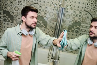 Günlük ev işleri sırasında günlük giysiler içinde elinde deterjan şişesi tutan ve banyodaki aynayı ıslak toz beziyle silen bir adam.
