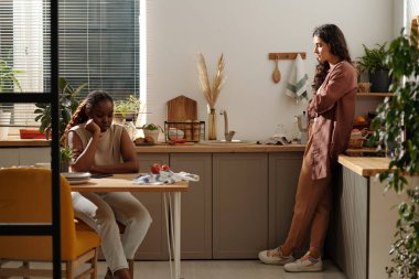 Kıskanç kadın mutfak tezgahının yanında dikilip akıllı telefonuyla kız arkadaşına bakarken suç ve yanlış anlaşılmaları dile getiriyor.