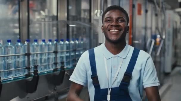 ミネラルウォーターボトリング工場にポーズしながら 青ポロシャツとジャンプスーツの折りたたみ手と笑顔で幸せなアフリカ系アメリカ人のエンジニアのミディアム刺し絵 — ストック動画