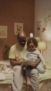 Afrika kökenli Amerikalı bir babanın küçük melez bir bebekle yatakta oturup animasyon film seçerken ya da akşam üzeri dekore edilmiş yatak odasında tabletten eğitim videosu izlerken tam bir dikey görüntüsü.