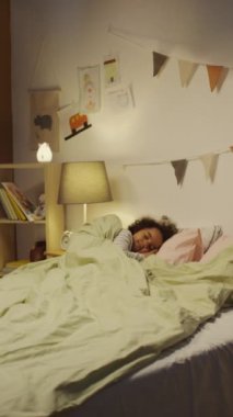 Küçük Afro-Amerikan bir çocuğun güzel dekore edilmiş, tahta oyuncaklar, resimler ve duvarlarda bayrak çelenkleriyle dolu bir odada çift kişilik bir yatakta uyuduğu dikey görüntü.