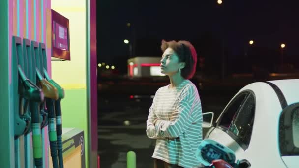 ガソリンポンプで白い近代車の近くに立っている若いアフリカ系アメリカ人ドライバーの中央側の映像は 夜に自動車が給油するのを待っています — ストック動画