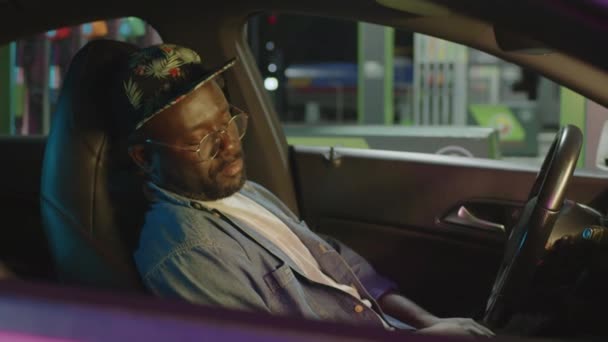 夜のガソリンスタンドで休憩中に車で眠っている若いアフリカ系アメリカ人の男の窓の映像を横切って — ストック動画