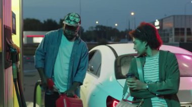 Benzin deposunu benzinle dolduran ve gece benzin istasyonunda kız arkadaşıyla konuşan Afro-Amerikan bir gencin orta boy görüntüleri.