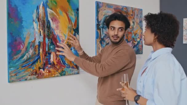 中东青年男子出席现代艺术展 与女伴站在表现主义绘画前 解释观点和作曲 女子聆听和点头的中景照片 — 图库视频影像