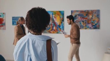 Orta ölçekli yakın plan çekimde, rastgele giyinmiş genç Afrikalı Amerikalı bir kadının müze koridorunda renkli soyut resimlerle duvara doğru yürümesi, onları durdurup yakından izlemesi.