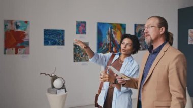 Orta boy olgun giyinmiş beyaz adam ve Afrikalı Amerikalı kadının müzedeki çağdaş sanat sergisine katıldığı, sanat eserlerini izlediği, tabloları tartıştığı ve şampanya yudumladığı bir fotoğraf.