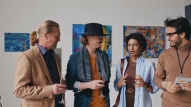 Çok ırklı modern sanat müzesi çalışanlarının orta boy portresi ve sergi açılışında tabloların önünde şampanya eşliğinde duran, sanat eserlerini tartışan, kameraya bakan ve gülümseyen konuklar.