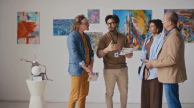 Kendine güvenen Arap erkek sanat galerisi çalışanı modern sanat sergisinde çeşitli ziyaretçilerle konuşuyor, soruları cevaplıyor, broşürlerdeki bilgilere ve sergilenen resimlere işaret ediyor.