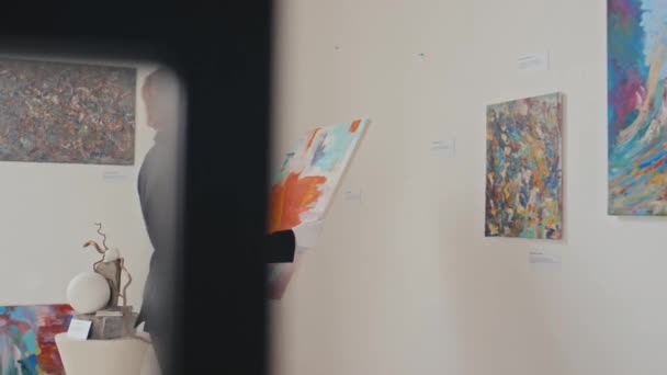 白い手袋の若い女性アートギャラリーの従業員のミディアムフルショットは カラフルな抽象絵画を運び 慎重に壁に掛け 新しい展覧会のために準備しながら — ストック動画