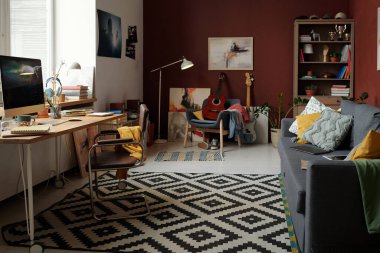 Geniş oturma odası, yerde halı, yastıklı rahat kanepe ve ofis müdürünün çalışma yeri arasında.