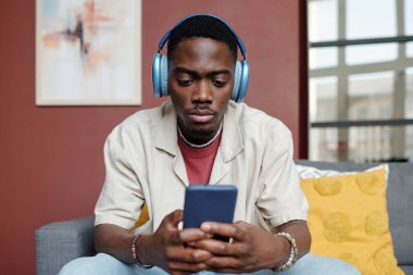 Kulaklıklı, akıllı telefon ekranına bakan ve evde kameranın önünde oturup online video izleyen genç, ciddi bir adam.