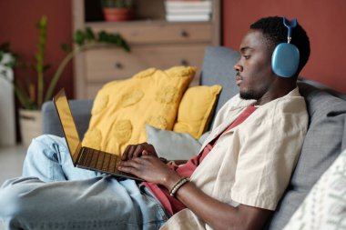 Genç iş adamı ya da blogcu kulaklıkla dizüstü bilgisayara bakıyor ve ağ sırasında oturma odasında otururken daktilo yazıyor.