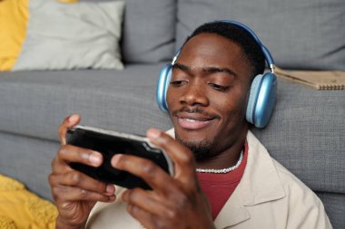 Kulaklıklı genç adam gri koltukta oturmuş oyun oynarken akıllı telefon ekranına bakıyor.