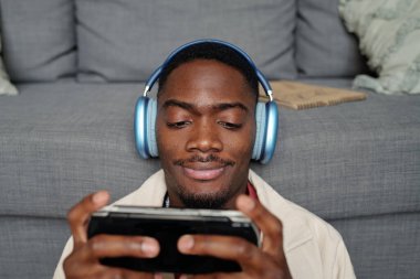 Kulaklık takan genç bir erkek oyuncu kanepenin karşısında oturuyor ve oyun sırasında oyun alanındaki akıllı telefon ekranına bakıyor.